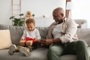 Black Senior Grandfather Giving Birthday Present To Grandson At Home