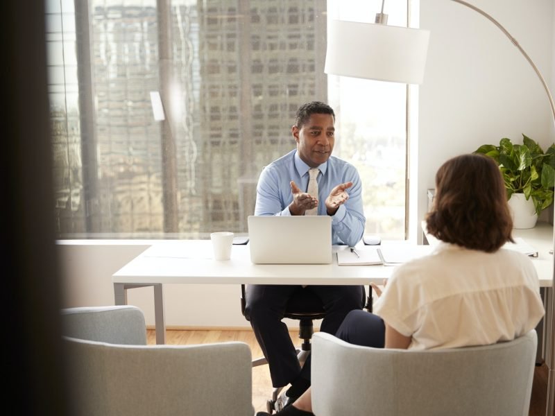 Male Financial Advisor In Modern Office Sitting At Desk Meeting Female Client